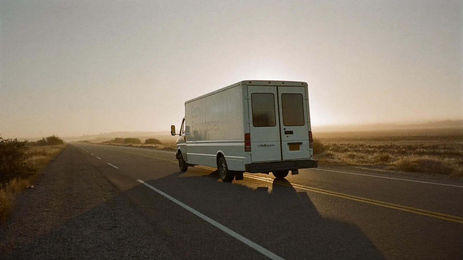 A plain white food truck driving down a two-lane highway toward the sunrise.