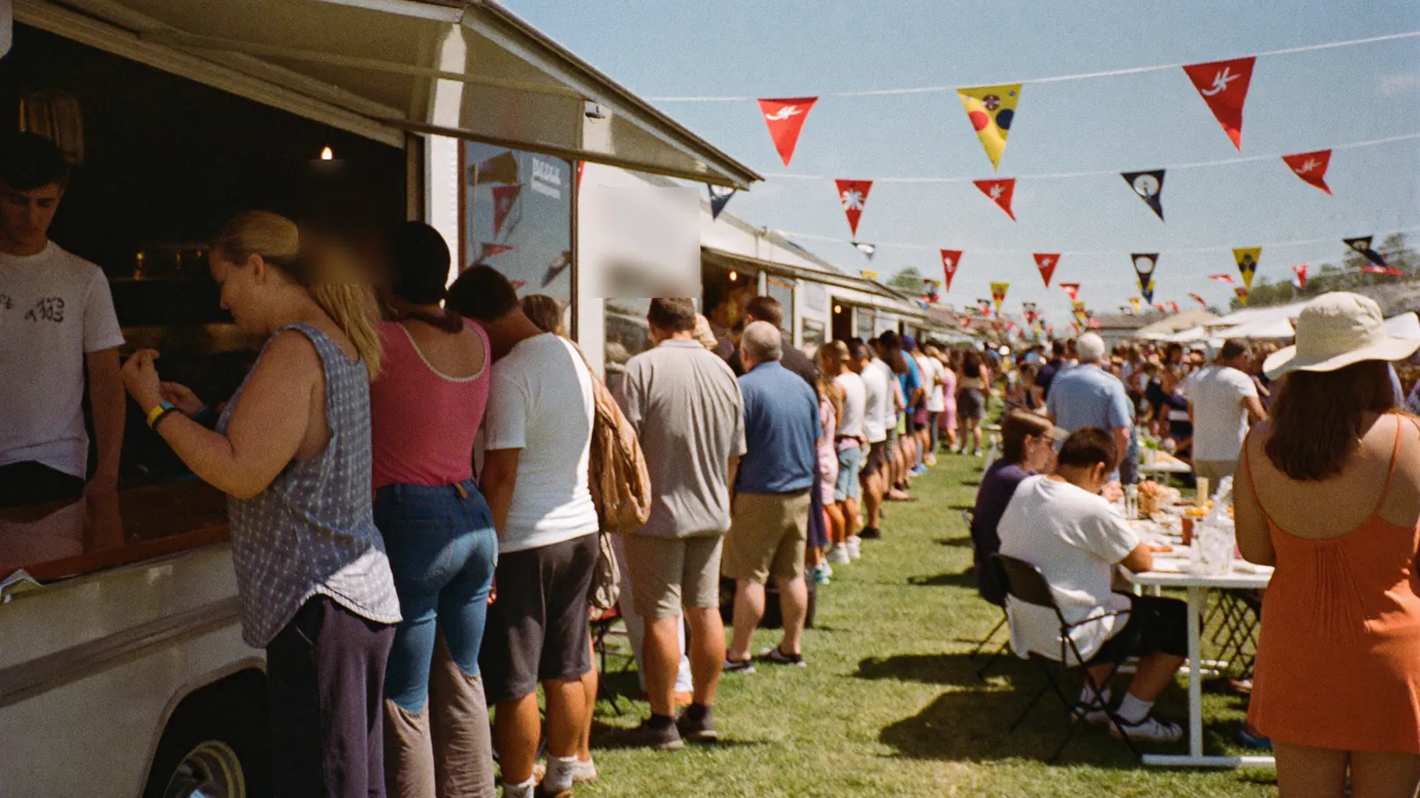 A long line of people at a food truck window at a sunny street festival, bunting overhead.