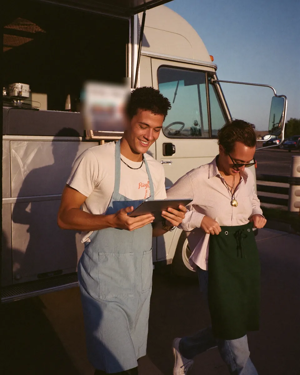 Two crew members in aprons arriving at a food truck at golden hour, one tapping a tablet by the back door.