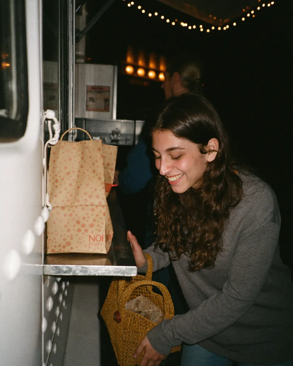 A customer smiling as she takes a paper bag from a food truck window at night.